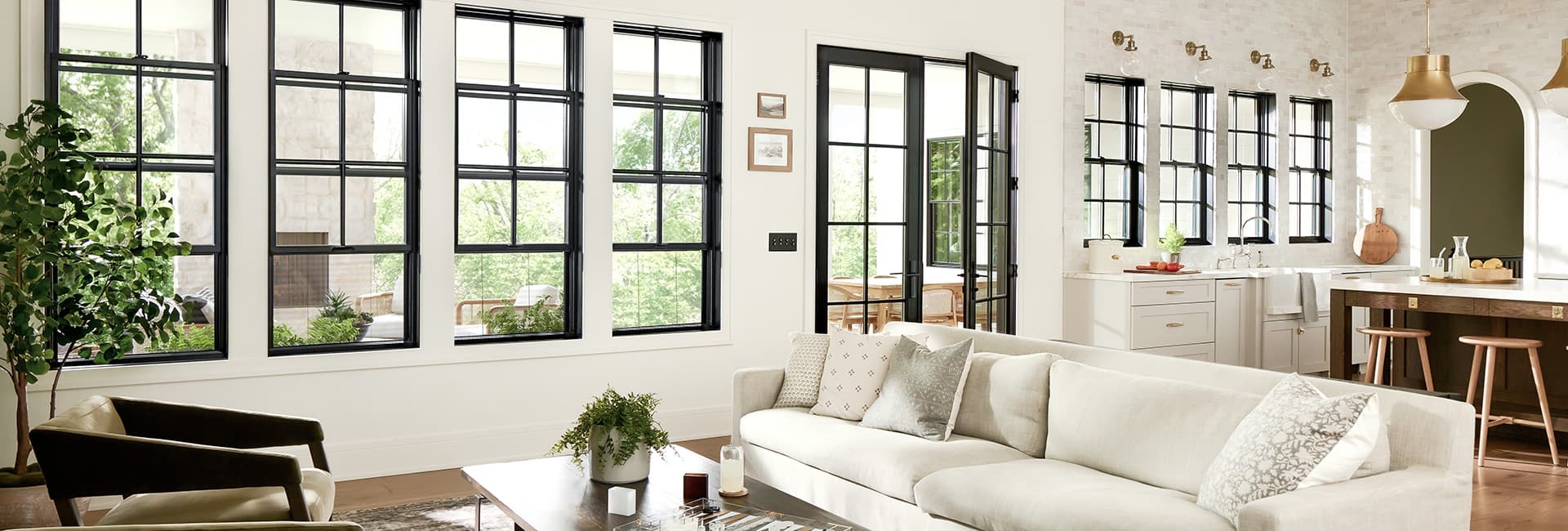 Bright open-concept living room and kitchen with large black-framed windows, neutral sofa, wood island with stools, brass pendant lights and indoor plants.