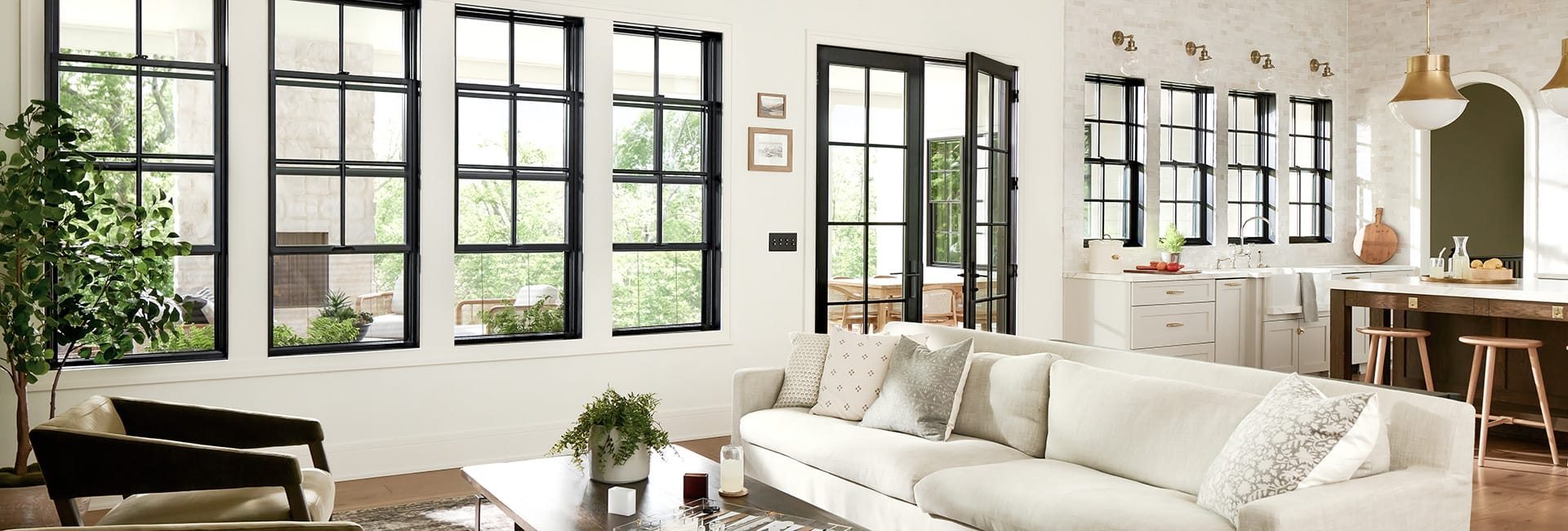 Bright open-concept living room and kitchen with large black-framed windows, neutral sofa, wood island with stools, brass pendant lights and indoor plants.