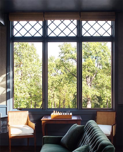Elegant home office with dark walls and large Pella fixed grid windows overlooking green trees