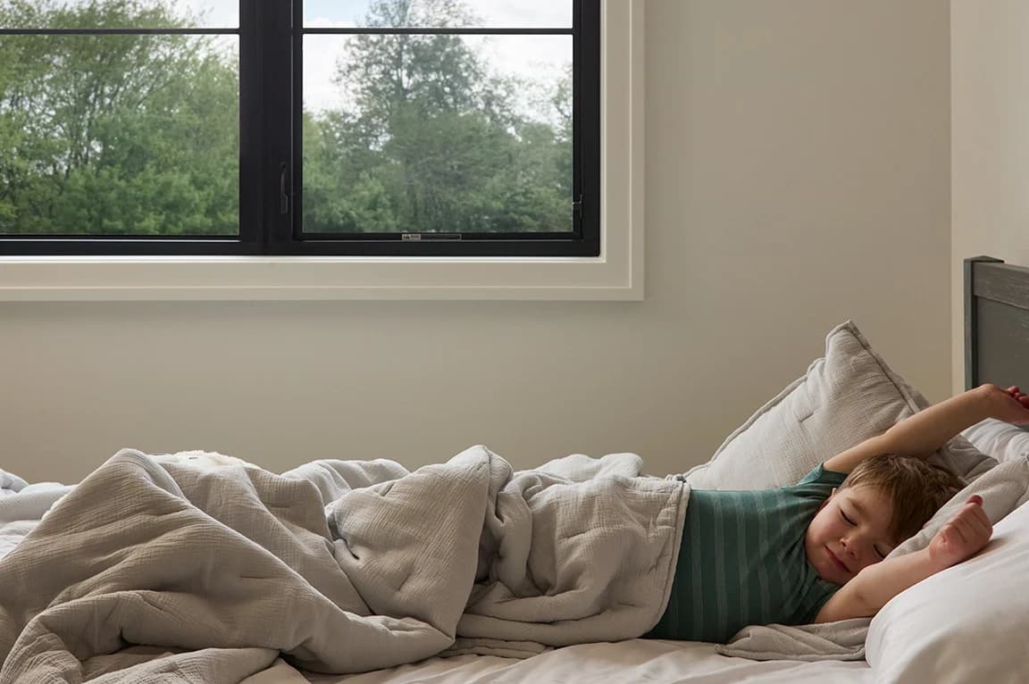 A child sleeps peacefully in a bed near a large soundproof black-framed window with trees outside.