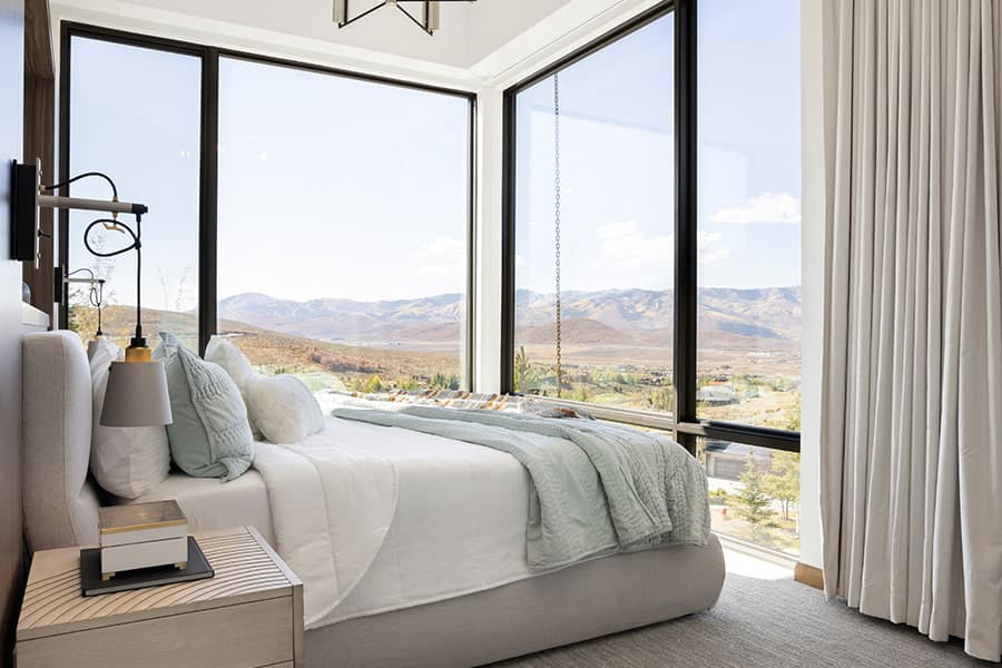 a large bed in the corner of floor-to-ceiling windows with large drapes.