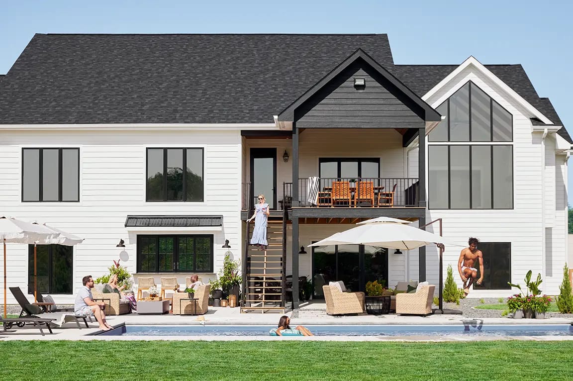 Two-story modern white farmhouse with black roof, backyard pool, patio seating and people relaxing outdoors