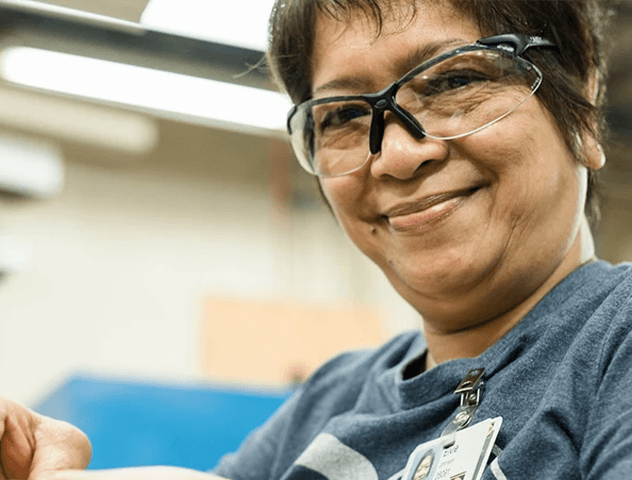 A woman wearing safety glasses and a name badge is smiling at the camera while working.