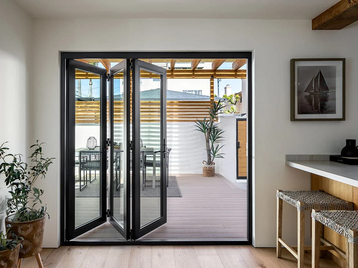 A black folding door opens to a modern patio with outdoor seating and potted plants.