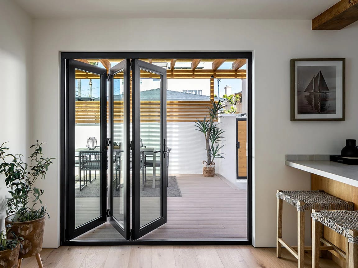 A black folding door opens to a modern patio with outdoor seating and potted plants.