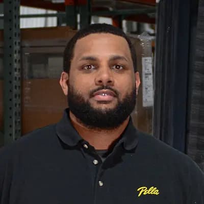 Employee wearing black Pella polo with blurred face standing in a warehouse aisle by metal shelving and stacked boxes