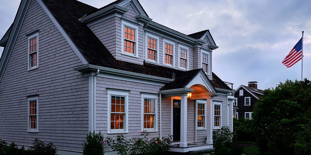 A cape cod-style home at dusk, featuring rows of white double-hung windows showing the lights inside the home.