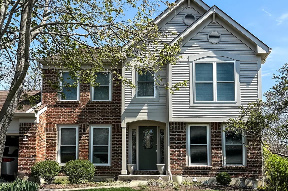 Two-story suburban house with red brick lower level and white siding gable, green front door, multiple windows with blinds and a landscaped front yard.