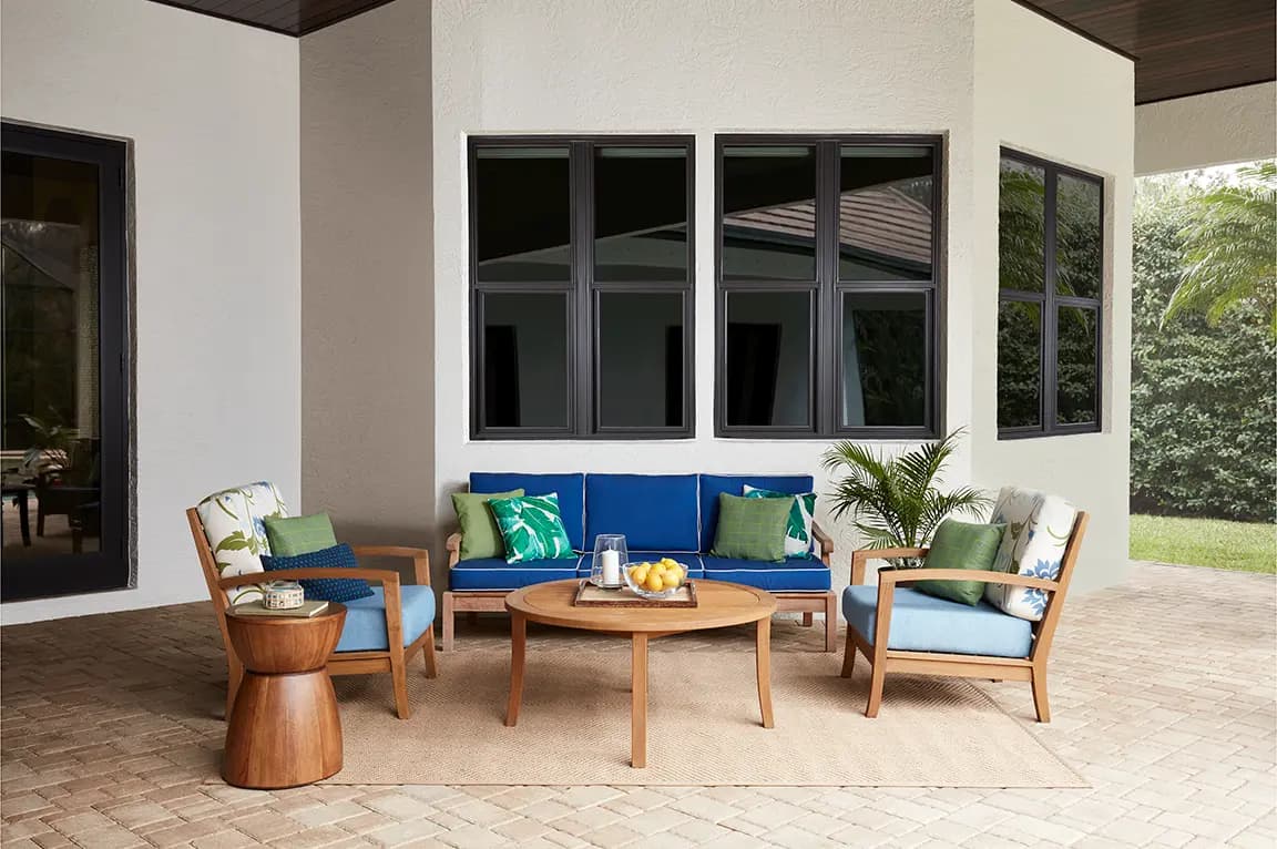 Outdoor patio furniture with blue-cushioned wooden sofa and armchairs, round wooden coffee table, tropical throw pillows, potted palm and windows.
