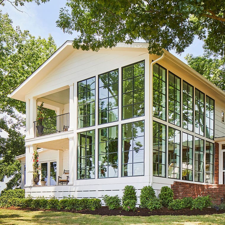 A modern house exterior features expansive floor-to-ceiling windows surrounded by greenery and trees.