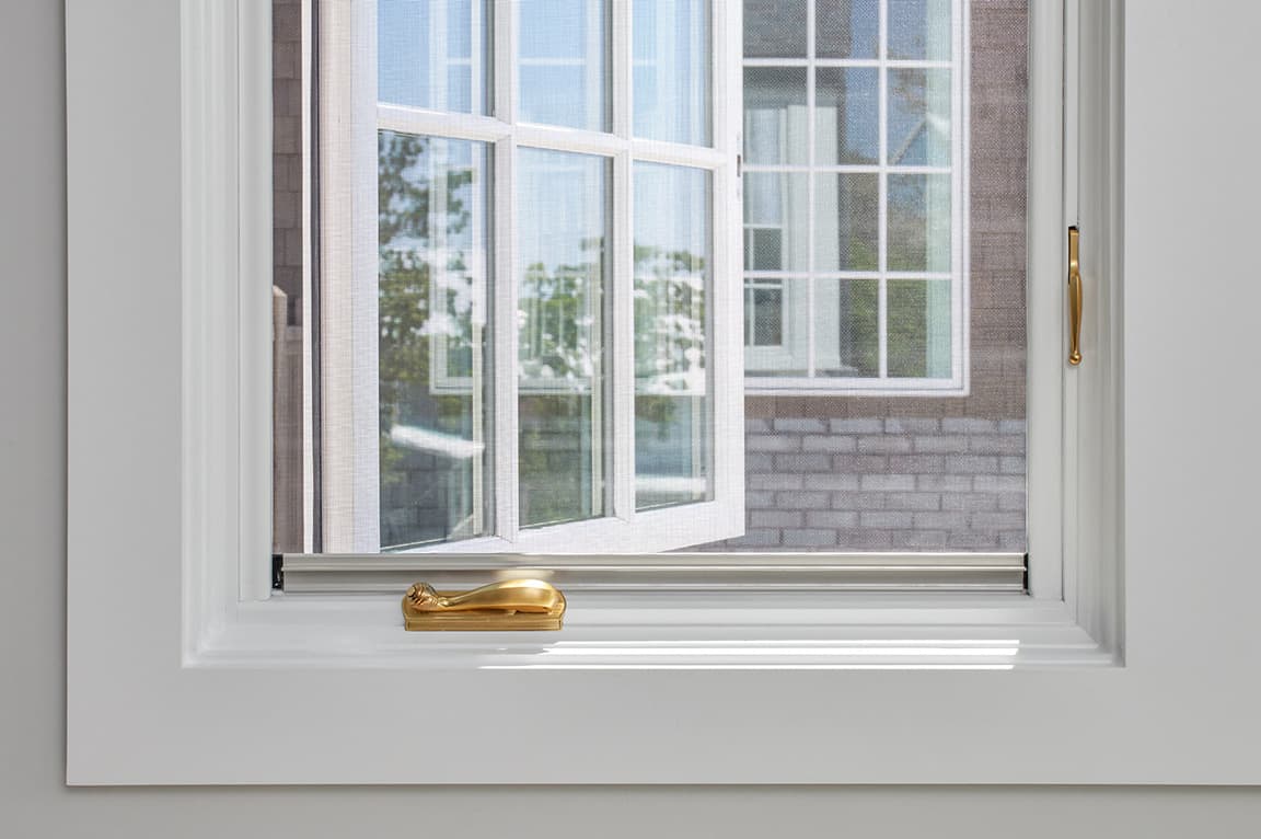 A close-up view of a casement window with a white frame and a gold handle, showcasing the outdoor scenery.