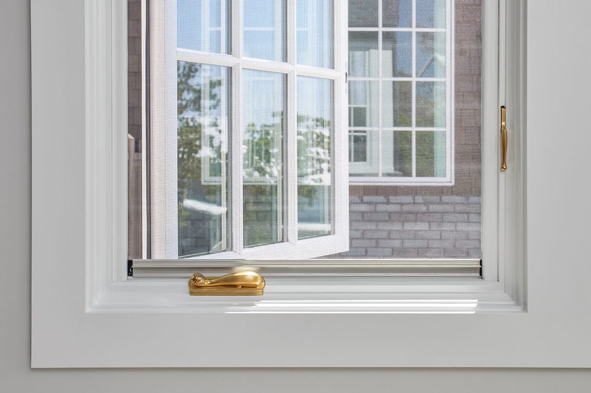 A close-up view of a casement window with a white frame and a gold handle, showcasing the outdoor scenery.