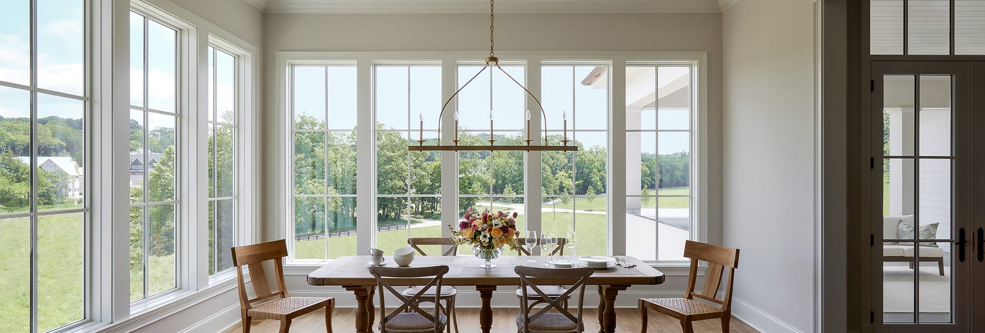 Sunlit dining room with large grid windows overlooking green landscape, wooden farmhouse table, chairs and floral centerpiece.