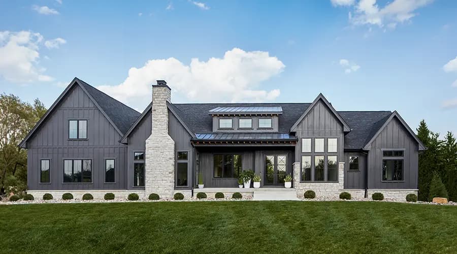 Modern farmhouse front facade with dark-gray board-and-batten siding, stone chimney and foundation, large windows and manicured lawn