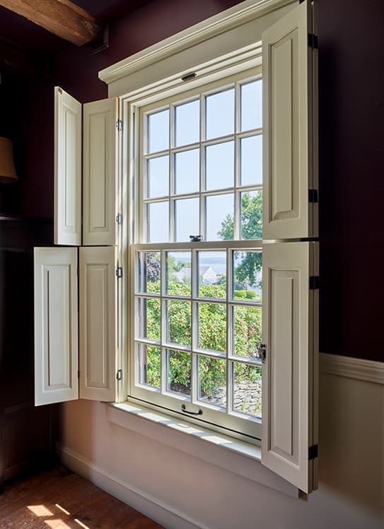 A large double-hung window painted in an off-white color has traditional grilles and folding blinds on both sides of the window.