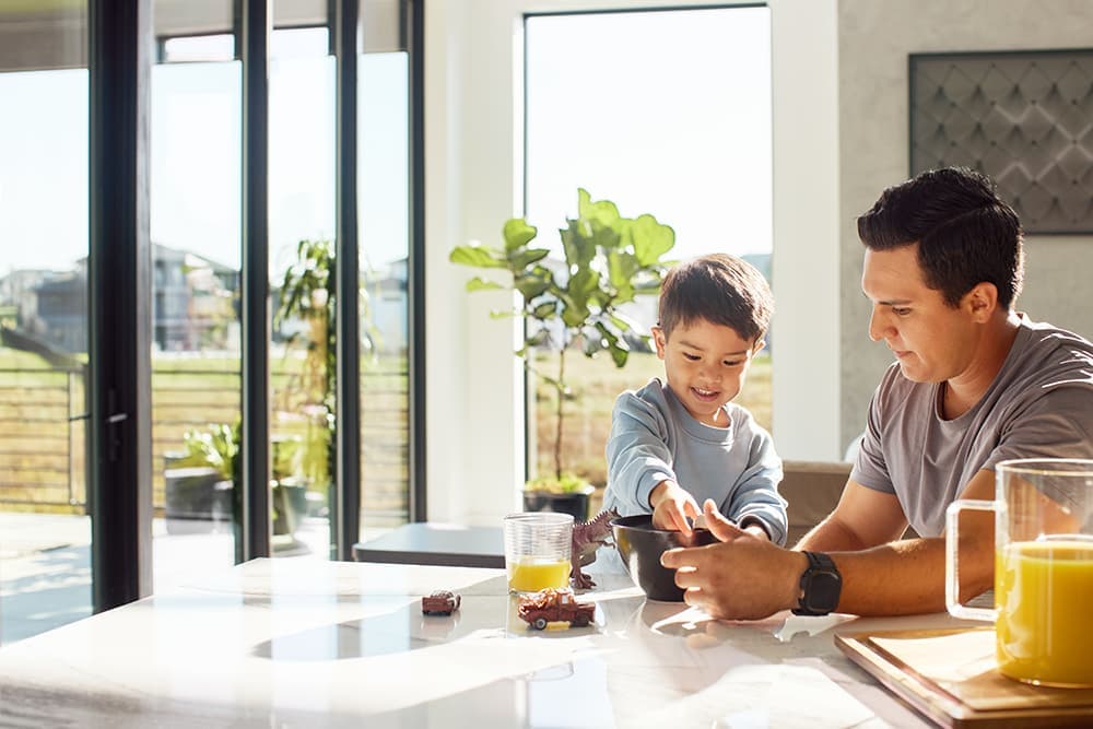A father and son are playing at the kitchen table with large floor-to-ceiling window on two walls.