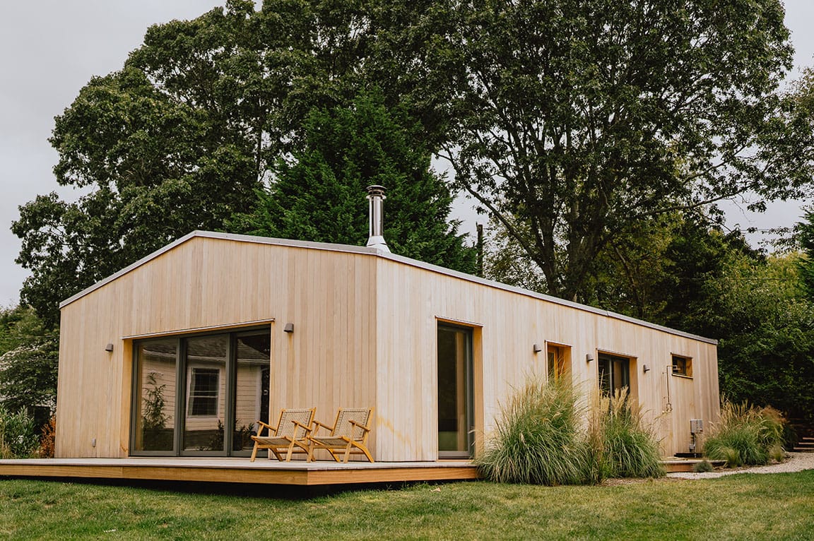 A modern wood-clad house with large energy efficient windows and sliding doors, surrounded by greenery.