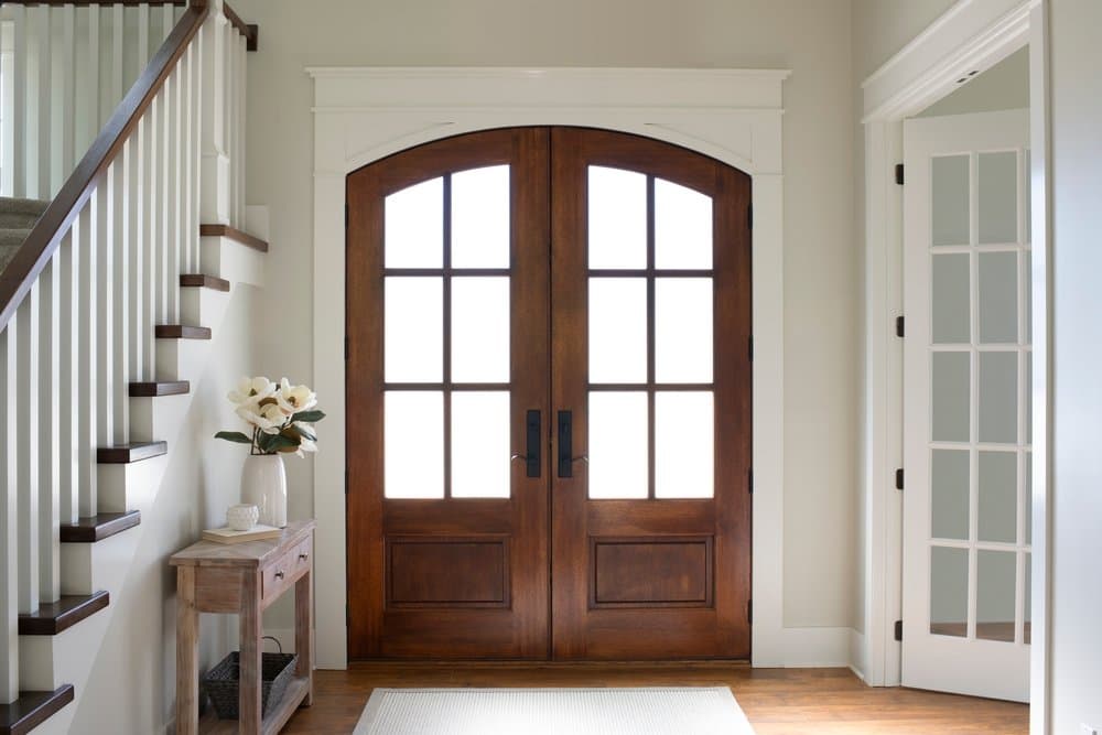 Wood Double Door Provides Warm Contrast To White Entryway White interior entryway with a wood double door with an arched top and traditional grilles on the 3/4 glass