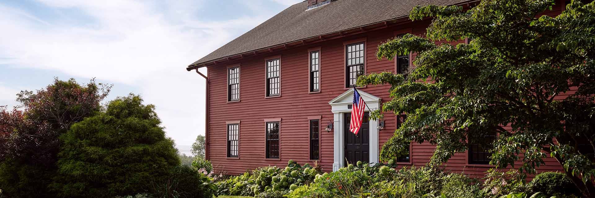 Red two-story colonial house with white trim, black front door and American flag, surrounded by lush garden and trees