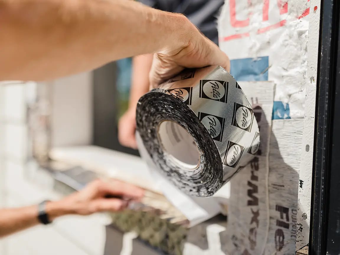 Roll of self-adhesive window flashing tape being applied to house wrap around a window opening during exterior construction