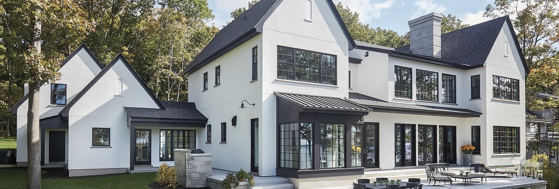 Modern white two-story luxury home with black-framed windows, gabled roof and outdoor patio on a wooded lot