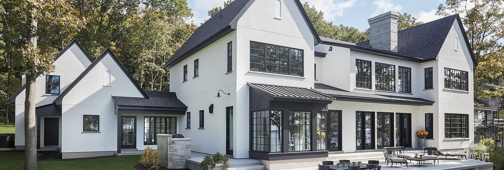 Modern white two-story luxury home with black-framed windows, gabled roof and outdoor patio on a wooded lot