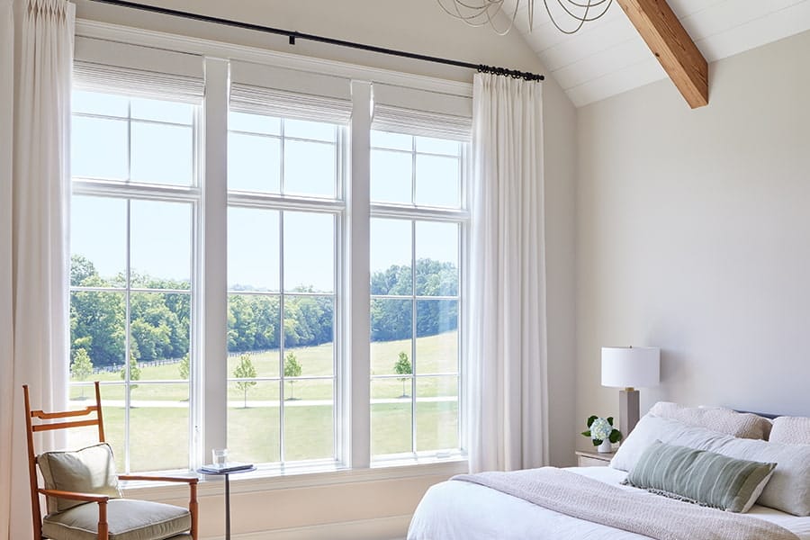 Bright bedroom with large grid-pane windows and white curtains, vaulted ceiling with wood beam, bed with green pillow and bedside lamp overlooking a sunny green field.