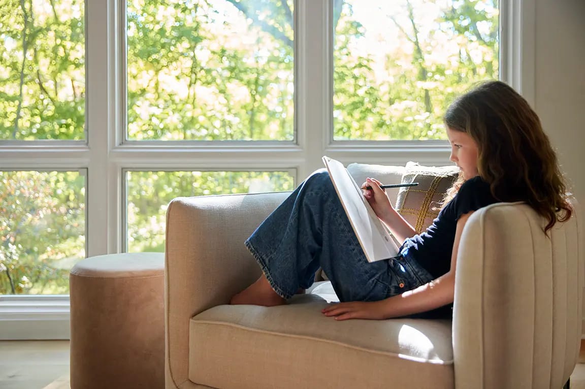 Young child sketching in a sketchbook while lounging on a beige sofa beside a large sunlit window in a cozy living room